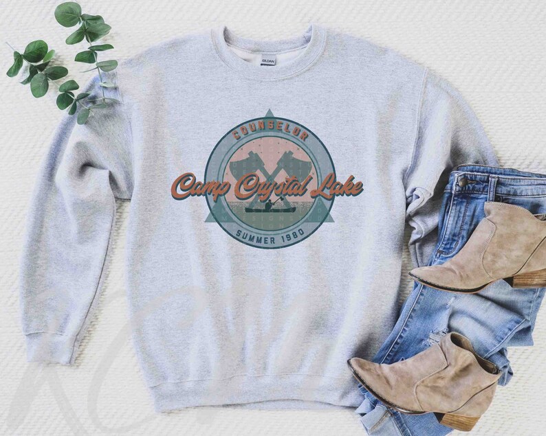 Op de afbeelding: Grijs sweatshirt met een vintage-stijl grafische afbeelding van een kamplogo. Het logo heeft een gekruist bijlontwerp, de tekst "Camp Crystal Lake" en de woorden "Counselor Summer 1980".