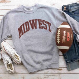 May include: Gray sweatshirt with the word "MIDWEST" in maroon letters. The sweatshirt is paired with blue jeans, white sneakers, and a brown football.
