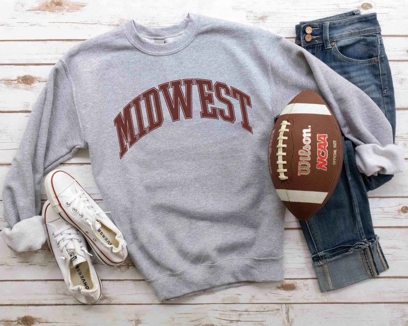 May include: Gray sweatshirt with the word "MIDWEST" in maroon letters. The sweatshirt is paired with blue jeans, white sneakers, and a brown football.