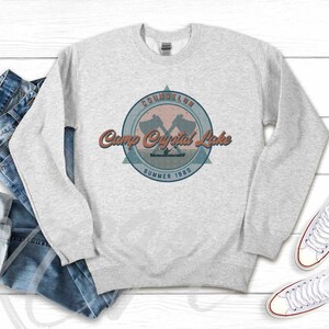 Op de afbeelding: Grijs sweatshirt met een vintage-stijl kampeerlogo. Het logo heeft een gekruist bijlontwerp en de tekst "COUNSELOR Camp Crystal Lake SUMMER 1980".