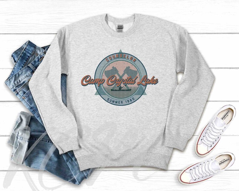 Op de afbeelding: Grijs sweatshirt met een vintage-stijl kampeerlogo. Het logo heeft een gekruist bijlontwerp en de tekst "COUNSELOR Camp Crystal Lake SUMMER 1980".