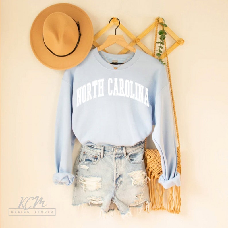 Op de afbeelding: Een lichtblauw sweatshirt met de tekst "NORTH CAROLINA" in witte letters. Het sweatshirt hangt aan een houten hanger met een rieten tas en een bruine hoed.