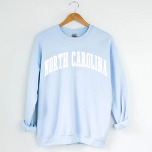Op de afbeelding: Een lichtblauw sweatshirt met de tekst "NORTH CAROLINA" in wit, in gebogen letters.