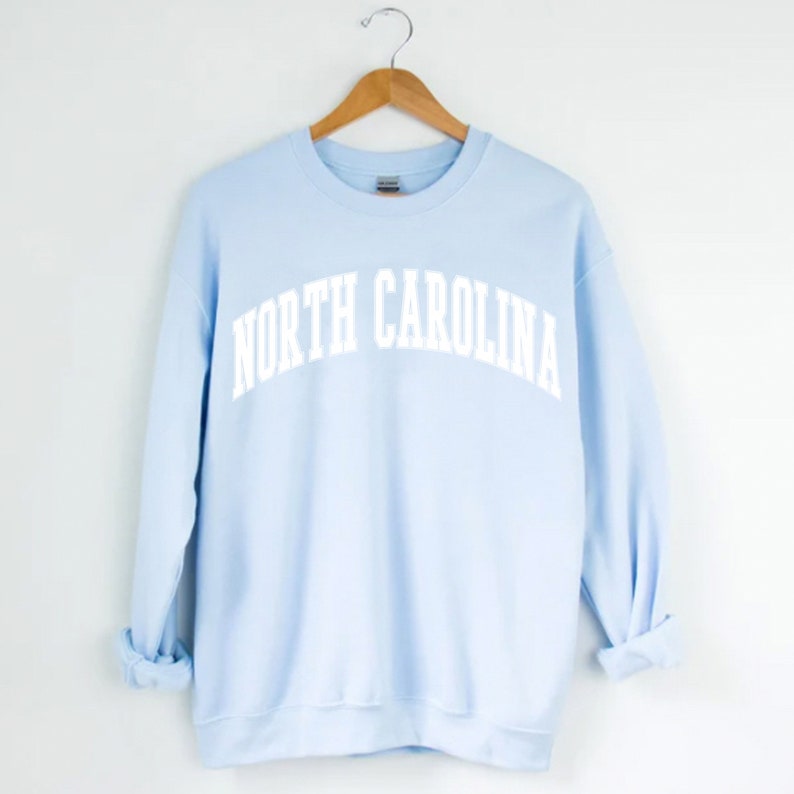 Op de afbeelding: Een lichtblauw sweatshirt met de tekst "NORTH CAROLINA" in wit, in gebogen letters.