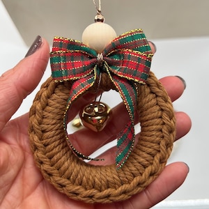 Op de afbeelding: Een bruin geweven kerstkransornament met een rood, groen en goud geruite strik. Een kleine gouden bel hangt in het midden. Het ornament heeft een houten kraal aan de bovenkant om op te hangen.