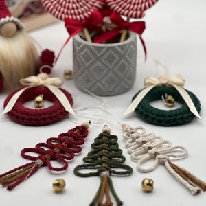 May include: Three handmade Christmas tree decorations made of yarn in red, green, and white. Each tree has a brown tassel and a wooden bead at the top. The decorations are displayed on a white surface with two gold bells.