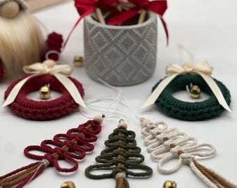 May include: Three handmade Christmas tree ornaments made of yarn in red, green, and white. Each tree has a brown tassel and a wooden bead at the top. The ornaments are displayed on a white surface with two gold bells.