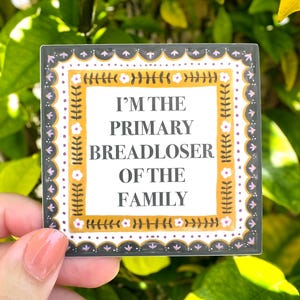 May include: A square sticker with a white center featuring the black text I'M THE PRIMARY BREADLOSER OF THE FAMILY. The text is framed by a mustard yellow border with a floral pattern, surrounded by a white border with small flowers and a dark gray edge.