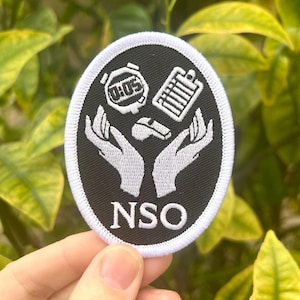 May include: White embroidered patch with a black background. The patch features two hands holding a stopwatch, a whistle, and a clipboard. The text "NSO" is written in white below the hands.