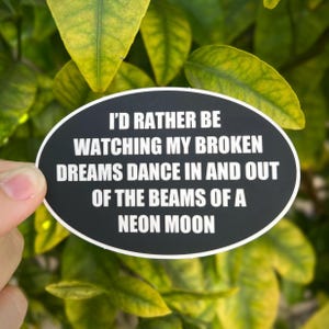 以下が含まれることがあります： ネオンムーンの光の中で、壊れた夢が踊るのを眺めている方が良い。という白文字が書かれた黒い楕円形のステッカーです。