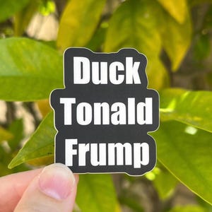 May include: A black and white sticker with the words "Duck Tonal Frump" in a bold, sans-serif font. The sticker is held in front of a background of green leaves.