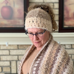 May include: A woman wearing a brown and white crocheted hat with a ponytail holder and a matching brown and white crocheted scarf. The scarf has a white and brown striped pattern.