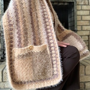 May include: A brown and white striped crocheted shawl with a pocket and a button. The shawl has a fuzzy texture and a white crocheted border.