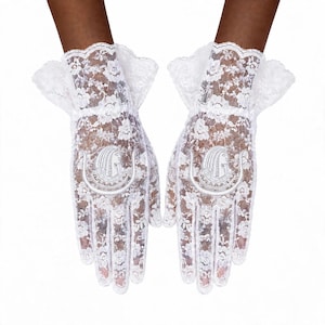May include: A pair of white lace gloves featuring a delicate floral pattern and a ruffled cuff. Each glove is adorned with a centered, embroidered white patch depicting the profile of an ancient Egyptian pharaoh in a traditional headdress.