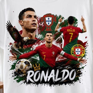 Può includere: Una t-shirt bianca a maniche corte con una stampa grafica di Cristiano Ronaldo con la maglia da calcio rossa e verde del Portogallo. Il design include un pallone, un'aquila, lo stemma nazionale portoghese e la scritta RONALDO in bianco.