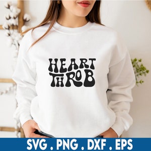 Op de afbeelding: Een witte sweatshirt met de woorden "HEART THROB" in een zwart, retro lettertype. De sweatshirt heeft een ronde hals en wordt door een persoon gedragen. Onderaan de afbeelding staan de bestandstypen: SVG, PNG, DXF en EPS.