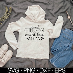 May include: A white hoodie with the text "grand children spoiled here" in black lettering and a small floral design below the text. The hoodie is laid out on a gray fabric background with a pair of blue jeans and a pair of white sneakers.
