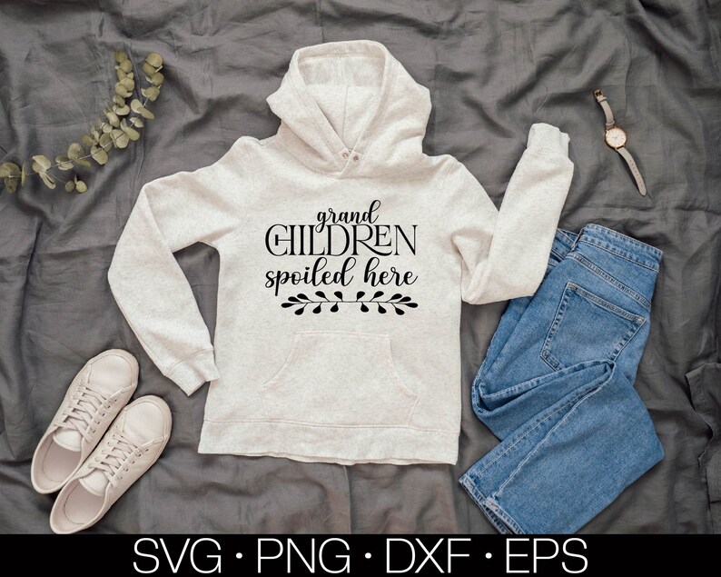May include: A white hoodie with the text "grand children spoiled here" in black lettering and a small floral design below the text. The hoodie is laid out on a gray fabric background with a pair of blue jeans and a pair of white sneakers.