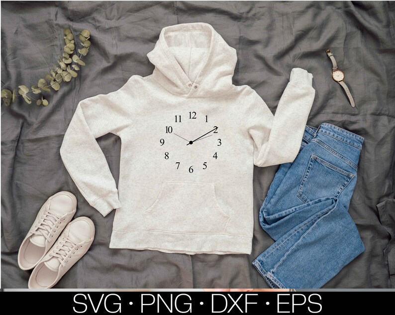 May include: A light grey hooded sweatshirt with a clock face design on the front. The clock face has black hands and black numbers. The sweatshirt is lying on a grey fabric background.