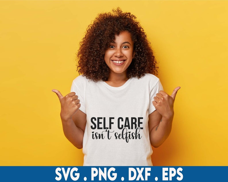 Puede incluir: Una mujer con el pelo rizado casta&ntilde;o sonr&iacute;e y se&ntilde;ala una camiseta blanca con el texto "SELF CARE isn't selfish" impreso en ella.