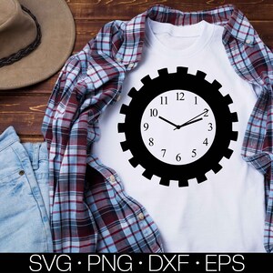 May include: A white t-shirt featuring a black and white gear-shaped clock design. The clock face has black numbers and hands. The t-shirt is laid out on a wooden surface with a plaid shirt and blue jeans.