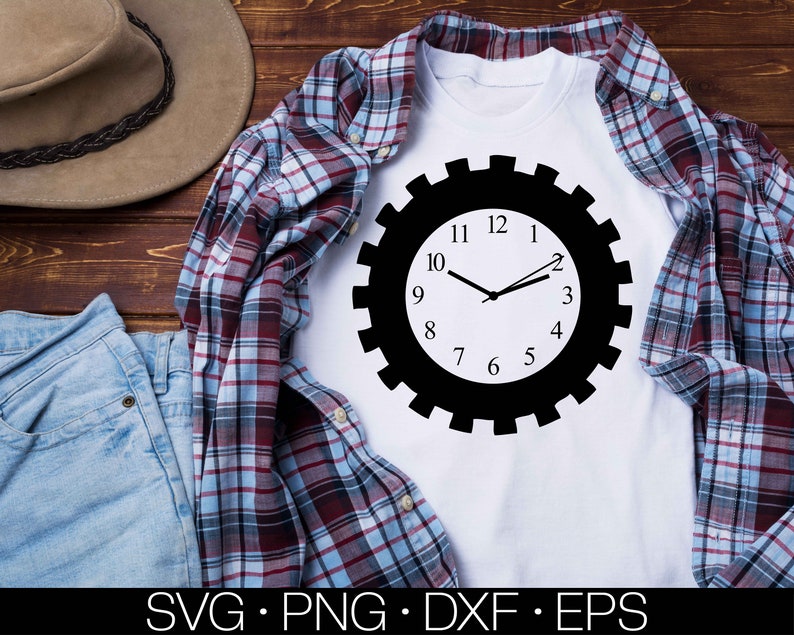 May include: A white t-shirt featuring a black and white gear-shaped clock design. The clock face has black numbers and hands. The t-shirt is laid out on a wooden surface with a plaid shirt and blue jeans.