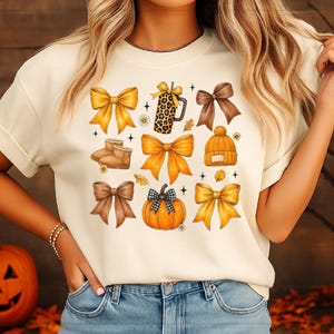 May include: Cream-colored t-shirt featuring a collage of autumn-themed illustrations. The design includes various bows in shades of yellow and brown, a pumpkin, a coffee mug, boots, and a beanie. The overall theme is fall and harvest.