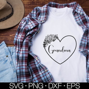 May include: White t-shirt with a black heart design and the word "Grandma" inside the heart. The heart is decorated with a floral design. The t-shirt is displayed on a plaid fabric background.