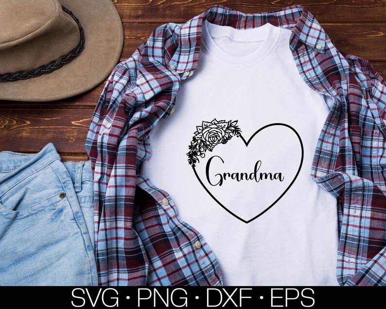 May include: White t-shirt with a black heart design and the word "Grandma" inside the heart. The heart is decorated with a floral design. The t-shirt is displayed on a plaid fabric background.