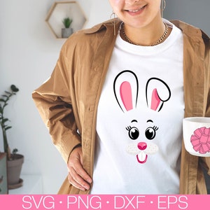 May include: A white t-shirt with a pink and black bunny face graphic. The bunny has large eyes, a pink nose, and a pink tongue sticking out. The t-shirt is being worn by a person with brown hair and a brown jacket.