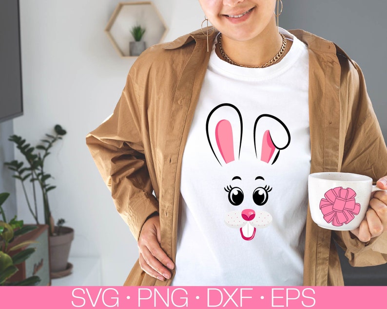 May include: A white t-shirt with a pink and black bunny face graphic. The bunny has large eyes, a pink nose, and a pink tongue sticking out. The t-shirt is being worn by a person with brown hair and a brown jacket.