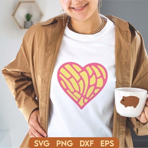 May include: A white t-shirt with a pink and yellow heart design. The heart is made up of a pattern of lines and swirls. The t-shirt is being worn by a person with a brown jacket.