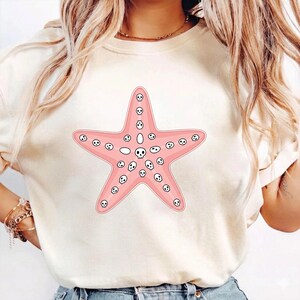 May include: A light-colored t-shirt featuring a pink starfish design. The starfish is covered in small white skull graphics. The design is centered on the shirt, creating a playful and unique aesthetic.