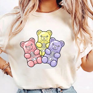 May include: A cream-colored t-shirt featuring three cartoon gummy bears in pink, yellow, and purple. Each bear has a skeleton design inside. The bears are outlined in black.
