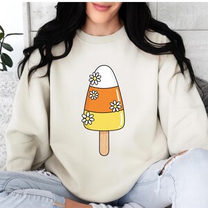 May include: A cream-colored sweatshirt featuring a candy corn popsicle design with white daisy accents. The popsicle has white, orange, and yellow sections, and is on a wooden stick. The design is centered on the front of the sweatshirt.