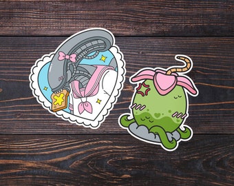 May include: Two cartoon stickers on a dark wood background. The first sticker is a heart-shaped frame with a grey alien wearing a pink and white sailor uniform and eating a piece of toast. The second sticker is a green alien with pink accents and a long, brown tail.