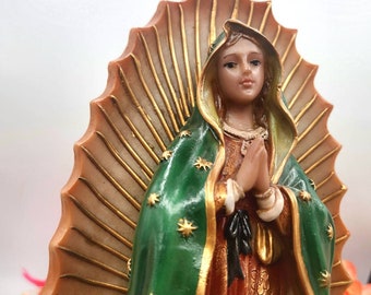May include: A religious figurine of the Virgin Mary, featuring a green cloak adorned with gold stars and a gold-trimmed robe. The statue has a halo with gold accents and is set against a white background. The figure's hands are clasped in prayer.