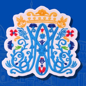 May include: Embroidered patch with a white scalloped edge, featuring a stylized monogram in light blue. The design includes a gold crown, red and blue flowers, and green leaves, set against a blue background with architectural sketches.