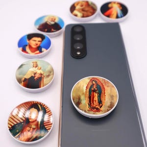 May include: A collection of round phone grips with different religious images. The grips are white with a clear plastic dome over the image. The images include a man with a beard, a man in a blue shirt, a woman with a baby, and a woman in a blue robe. The grips are on a white surface with a gray smartphone in the background.