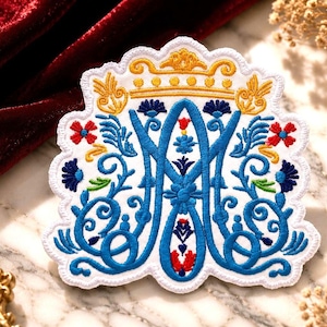 May include: An embroidered patch featuring a large blue monogram of the letters AM, topped with a yellow crown. The design includes intricate blue, red, and green floral and vine motifs on a white background with a scalloped white border edge.