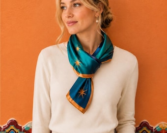 May include: A square silk scarf featuring a deep teal background with gold starburst patterns and a solid gold border. The scarf is tied around the neck, paired with a cream-colored long-sleeved sweater and matching cream-colored trousers.