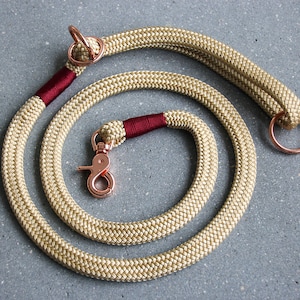 May include: A beige rope dog leash with a rose gold clip and two rose gold rings. The leash has a burgundy accent.