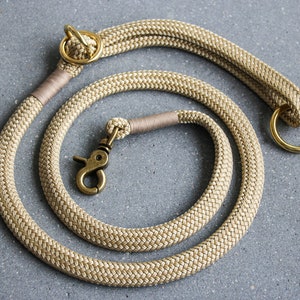 May include: Tan rope dog leash with brass hardware. The leash is coiled on a gray surface, showcasing its woven texture and the gold-colored metal clasp and rings. The leash is designed for pet owners.