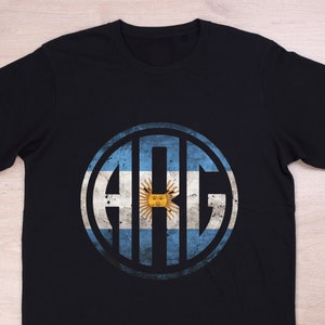 Puede incluir: Camiseta negra con un diseño circular que presenta las letras "ARG" en un círculo blanco con fondo azul y blanco. El círculo tiene un aspecto desgastado, vintage y las letras están en una fuente en negrita y de bloque. El fondo del círculo es de los colores de la bandera argentina.