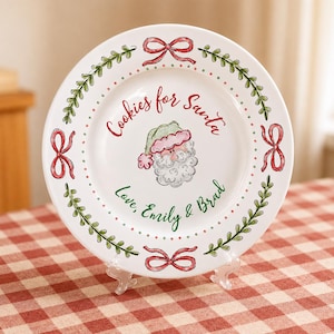 May include: A white ceramic plate featuring a hand-painted illustration of Santa Claus in the centre. The text "Cookies for Santa" appears above the illustration, and "Love, Emily & Brad" appears below. The rim is decorated with green vines and red bows.