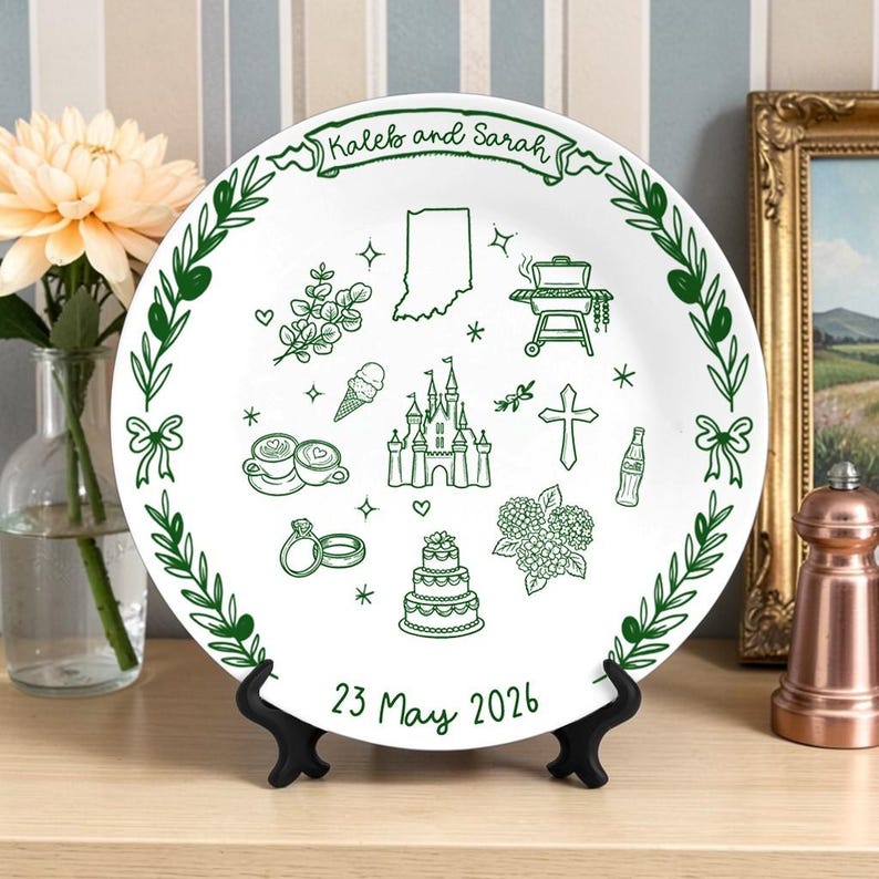 May include: A white ceramic plate featuring green line art illustrations including a castle, a wedding cake, rings, a cross, a grill, a map of Indiana, and coffee cups. The text reads Kaleb and Sarah at the top and 23 May 2026 at the bottom.