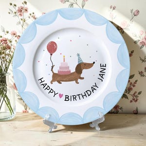 May include: A white ceramic plate with a scalloped light blue border featuring a brown dachshund wearing a party hat, carrying a pink tiered cake and a red balloon, with the text Happy Birthday Jane and a small pink heart printed in black.