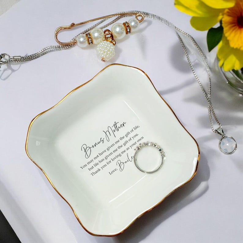 Op de afbeelding: Een witte keramische ringschaal met gouden rand, met de tekst "Bonus Mother". Een zilveren ring, een met parels versierde veiligheidsspeld en een zilveren ketting met hanger zijn ook aanwezig. De schaal bevat een ontroerende boodschap.