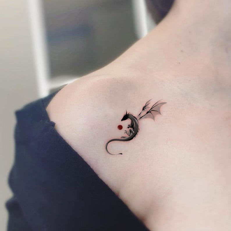 Pode incluir: Uma pequena tatuagem de drag&atilde;o preto no ombro de uma pessoa. O drag&atilde;o est&aacute; enroscado com as asas abertas. H&aacute; um pequeno ponto vermelho atr&aacute;s do drag&atilde;o.