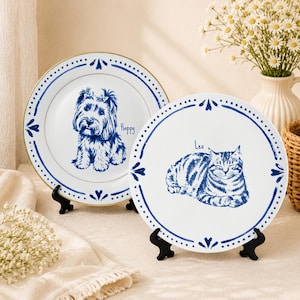 May include: Two white ceramic plates with blue decorative borders and gold rims. One plate features a blue illustration of a dog with the text Happy, while the other displays a blue illustration of a cat with the text Leo, both on black stands.